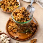 A jar of orange pistachio granola sitting on a small wooden plate.