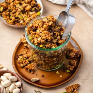 A jar of orange pistachio granola sitting on a small wooden plate.