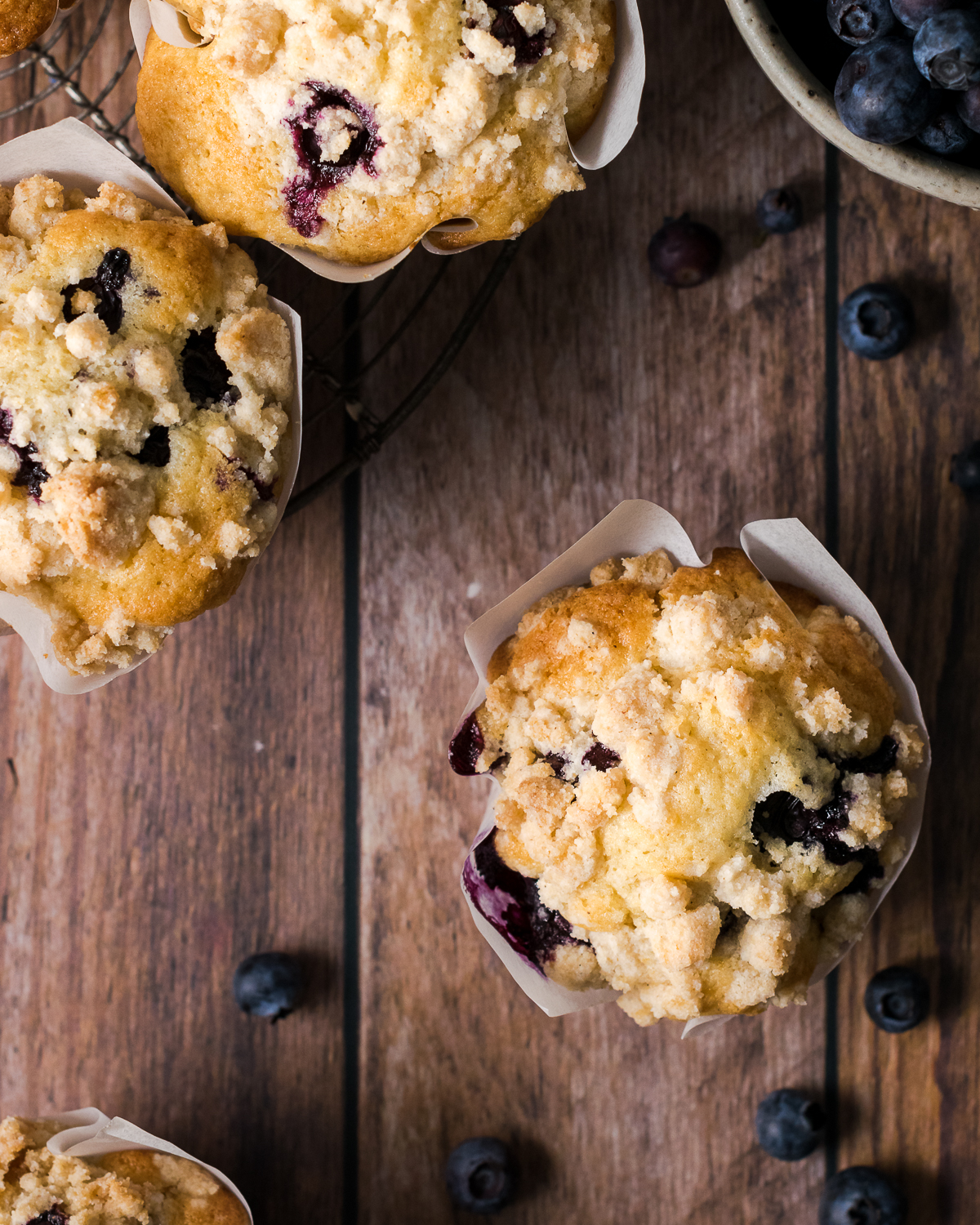 Three gluten free blueberry muffins sitting on a wooden table with a bowl of fresh blueberries.