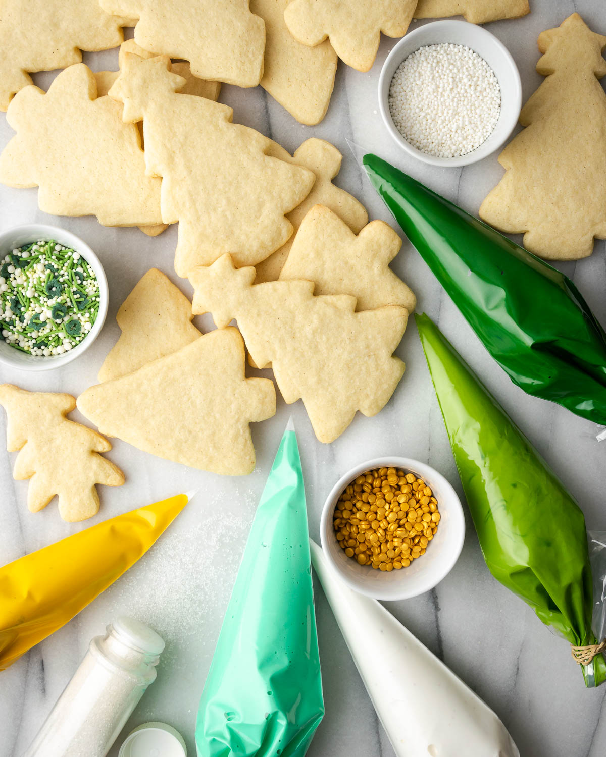 Gluten free sugar cookies cut into tree shapes and bags of royal icing laid out on a marble table.