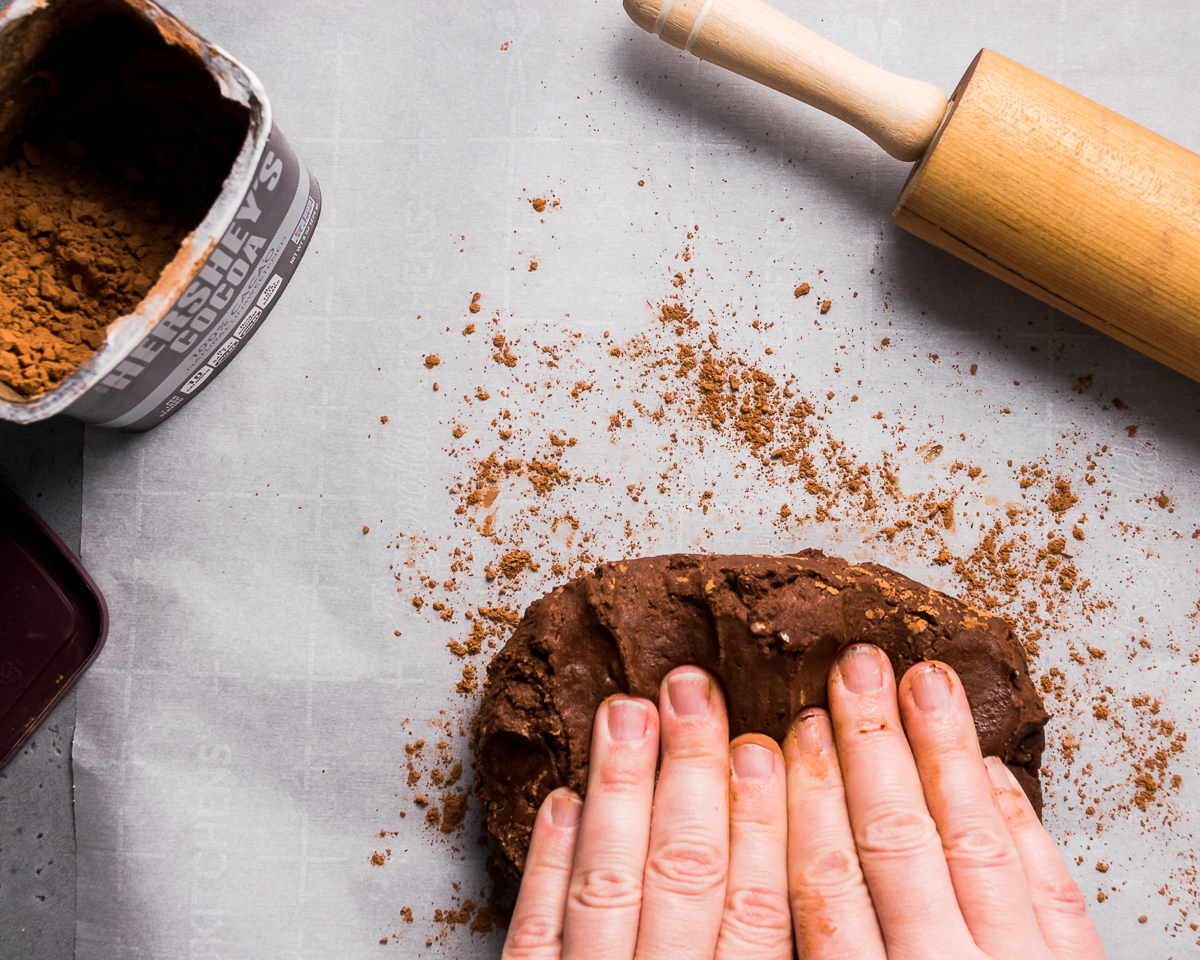 Hands kneading chocolate cookie dough on a piece of parchment paper.