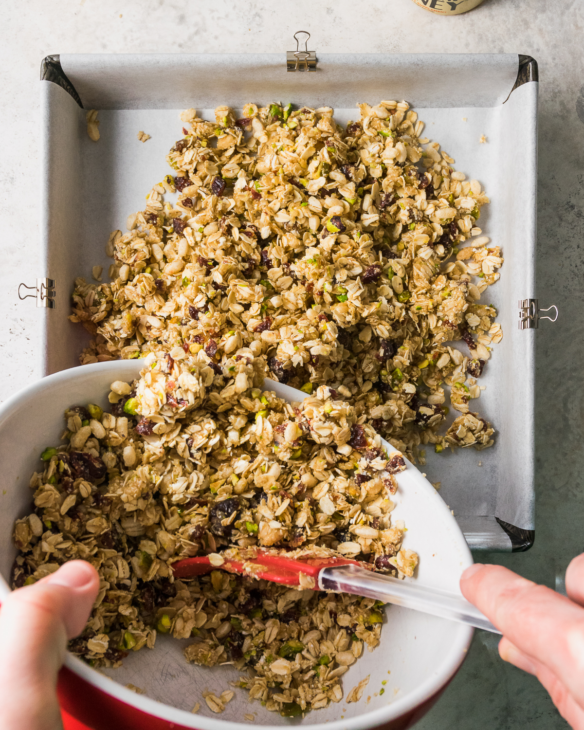 Mixed granola being poured into a square baking tin lined with paper.