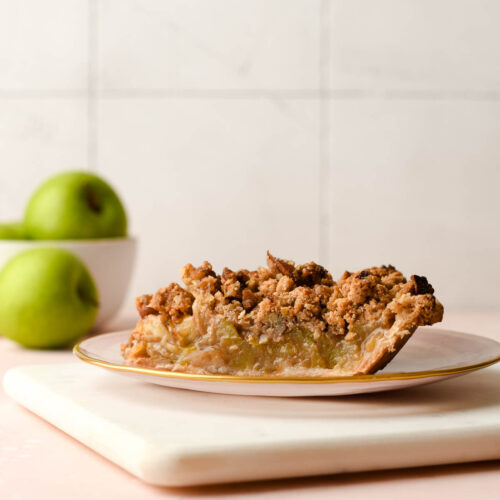 A slice of pie on a pink plate with green apples in the background.