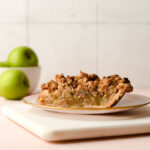 A slice of pie on a pink plate with green apples in the background.