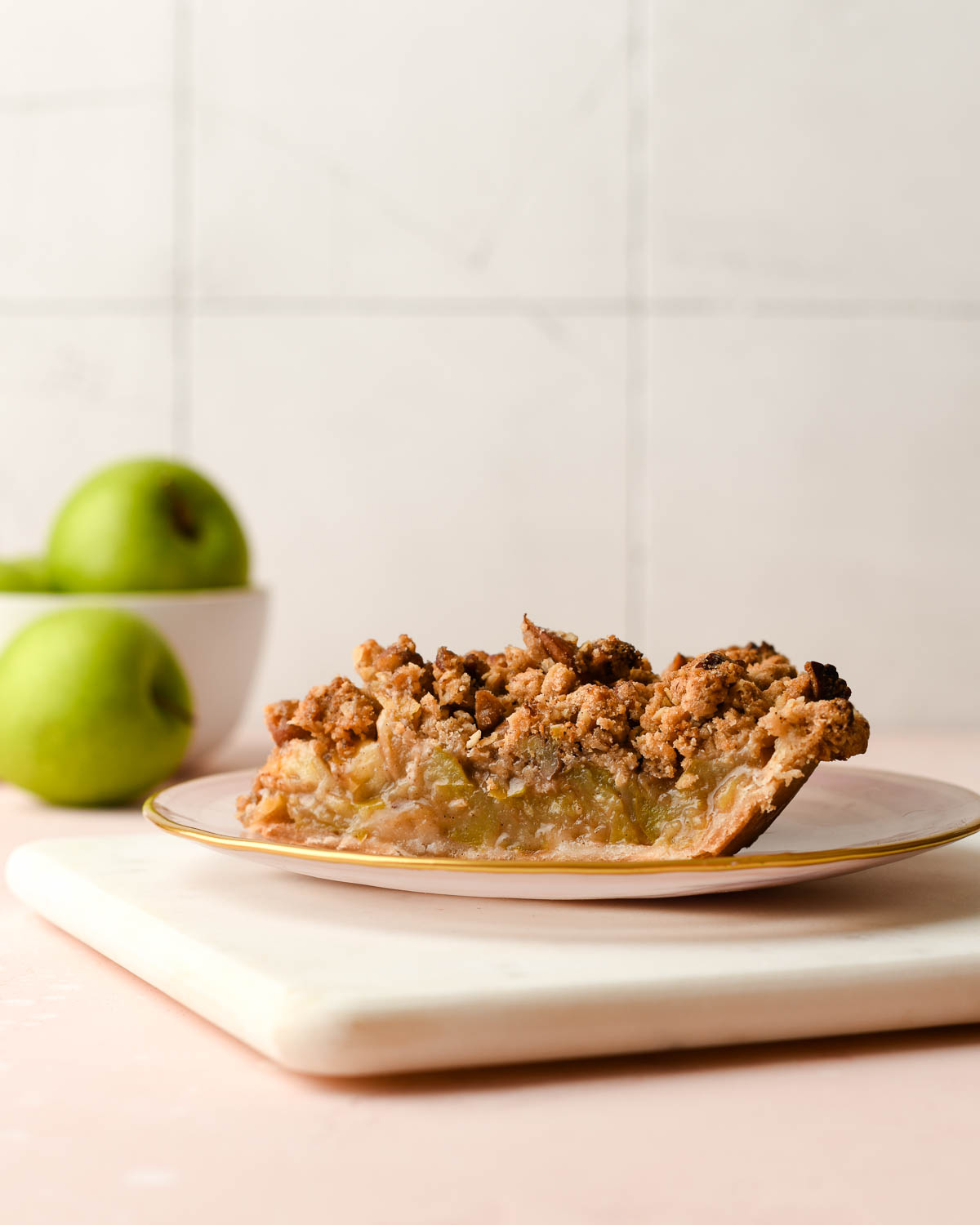 A slice of pie on a pink plate with green apples in the background.