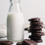A stack of thin mint cookies next to a jar of milk on a white table.
