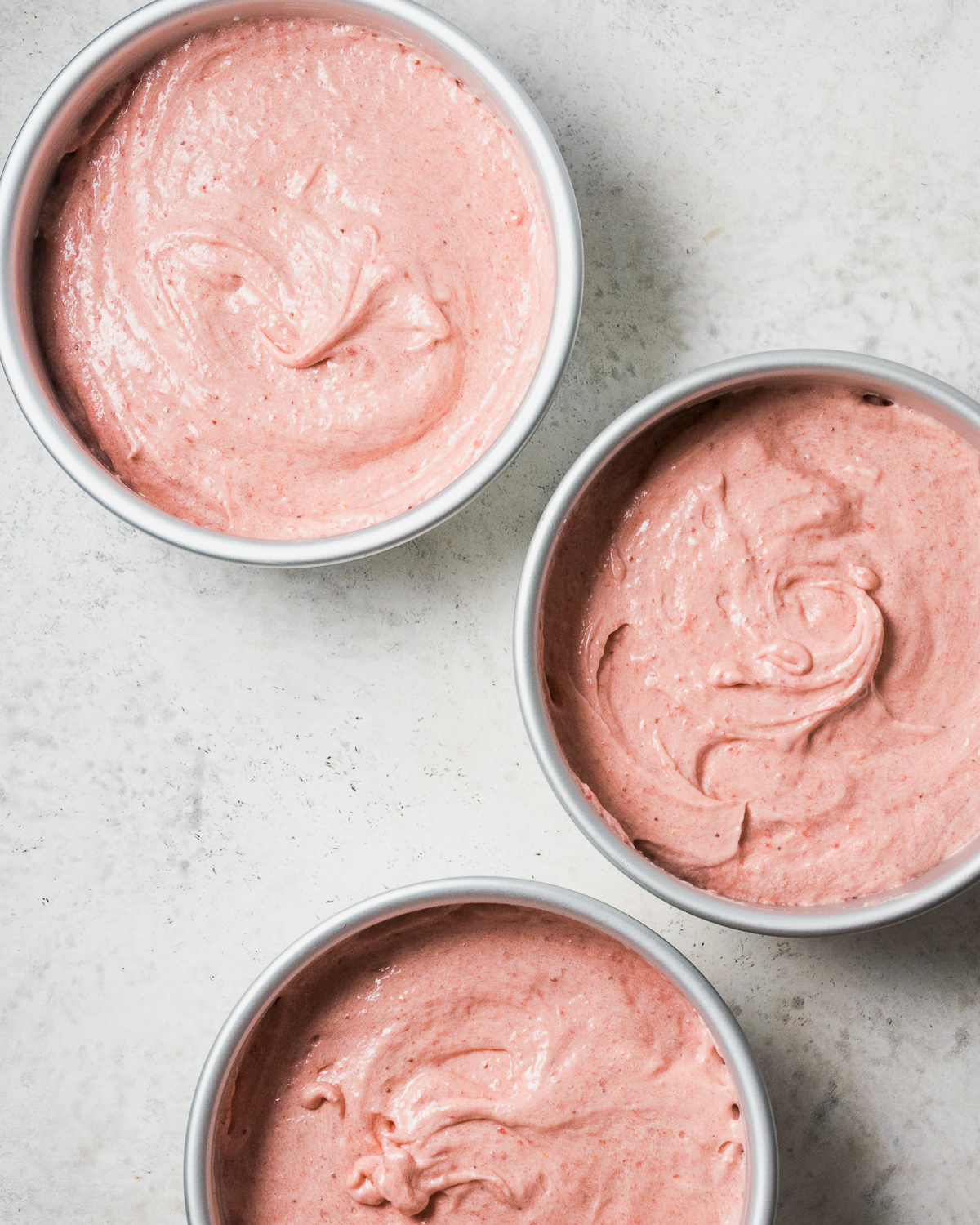 Three cake pans filled with pink batter on a white table.