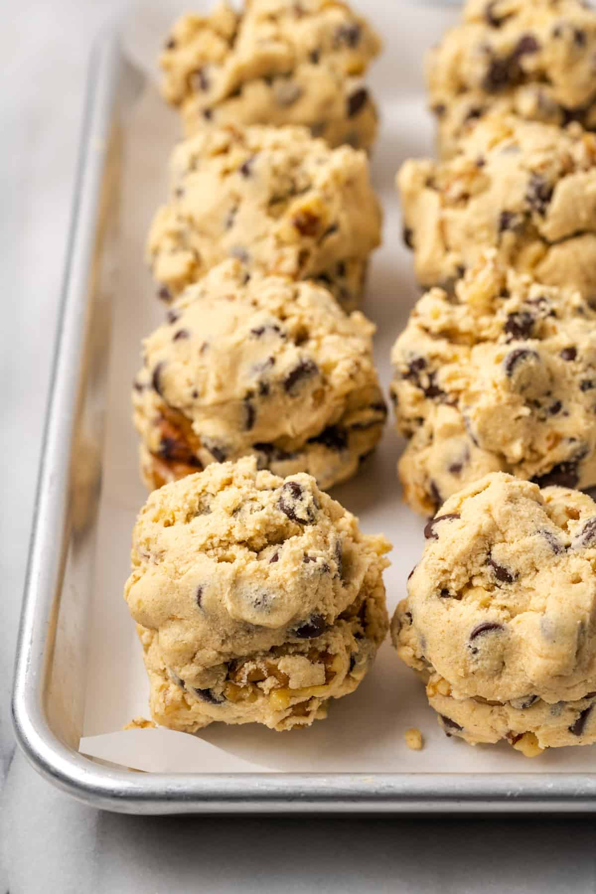 Large cookie dough balls laid in rows on a metal baking sheet.