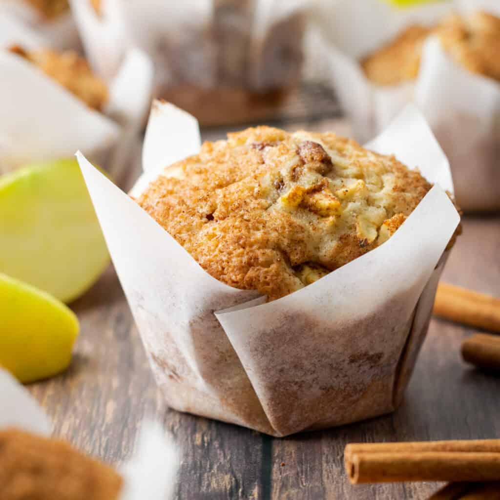 A gluten free apple muffin in a white paper wrapper sitting on a wooden table.