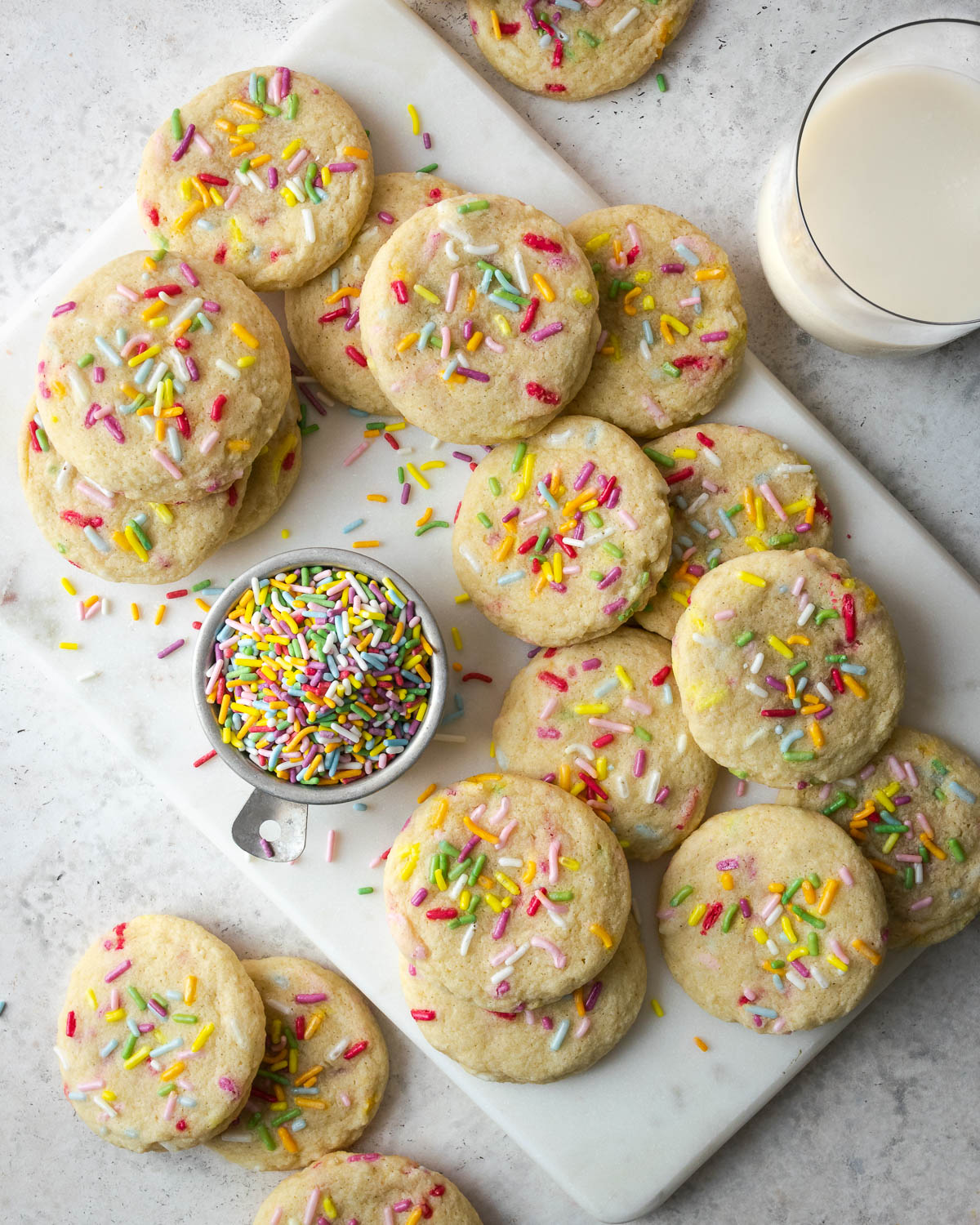 A batch of gluten free sugar cookies with sprinkles laying on a white cutting board with a glass of milk.