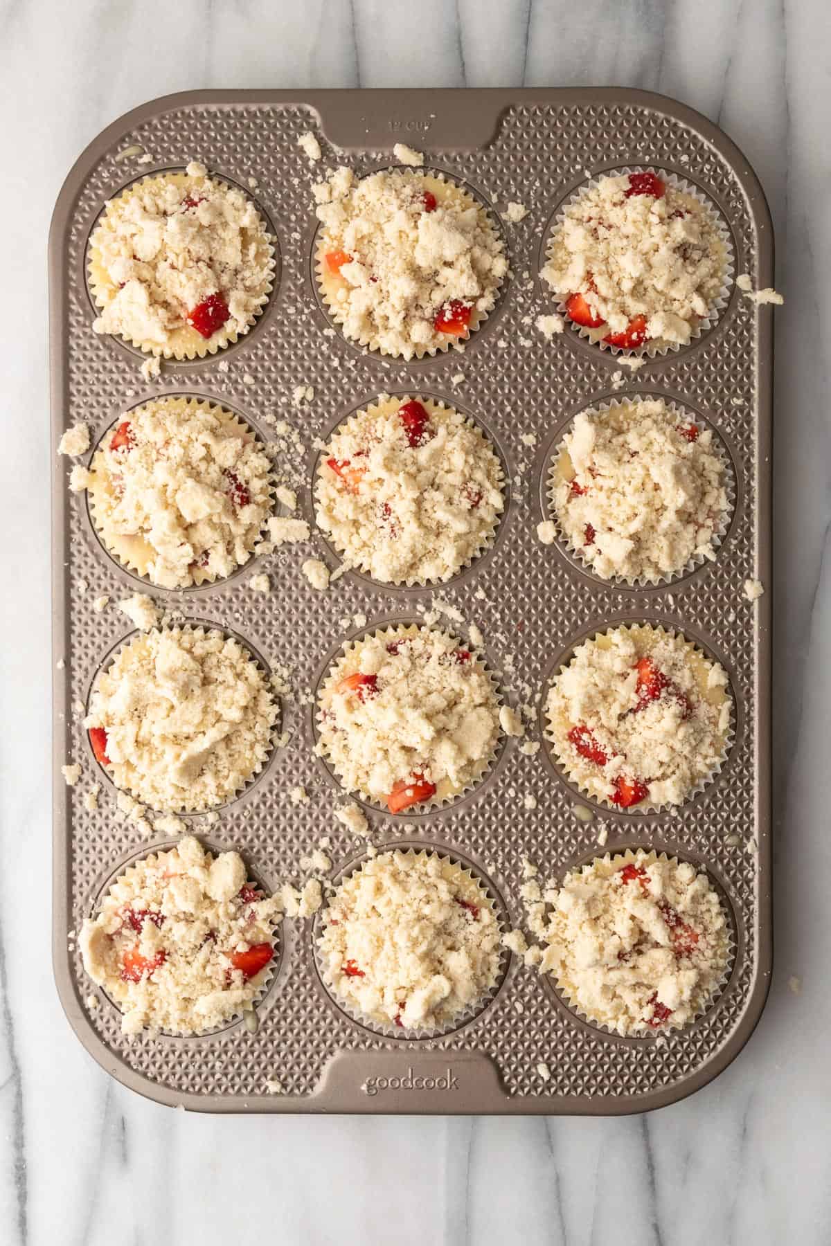 A muffin tin filled with unbaked strawberry muffin batter topped with crumble topping.
