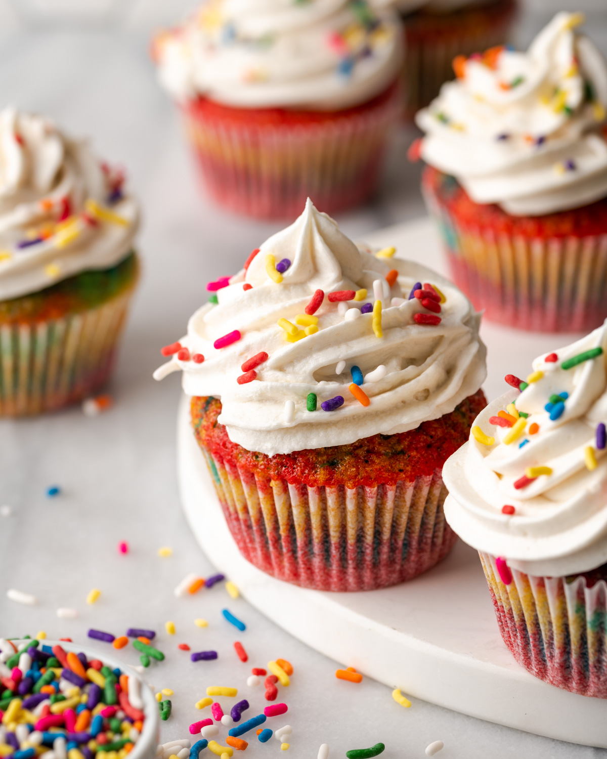 Tie dye cupcakes topped with vanilla buttercream and rainbow sprinkles sitting on a white table.