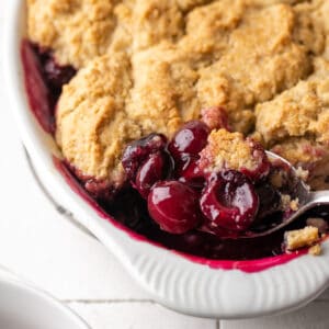 A scoop of cherry cobbler being lifted from a white baking dish.