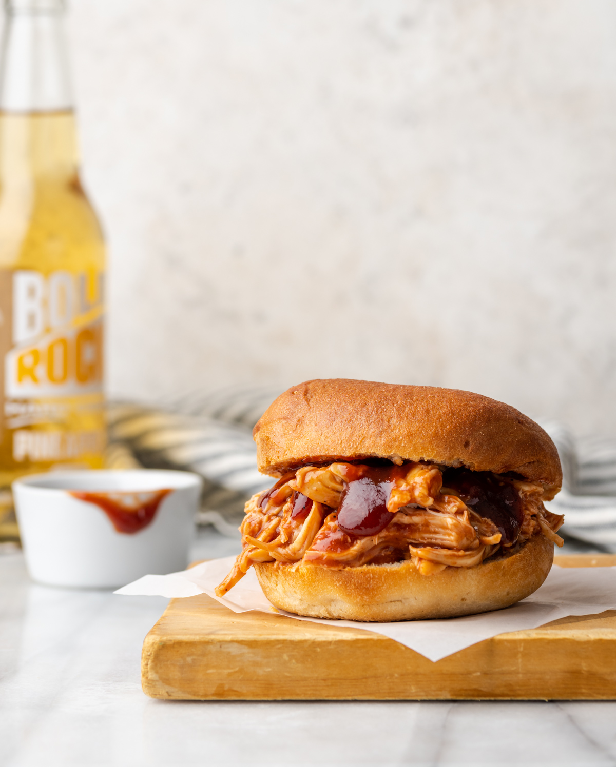 Pulled BBQ chicken sandwich on a cutting board with a drink in the background.