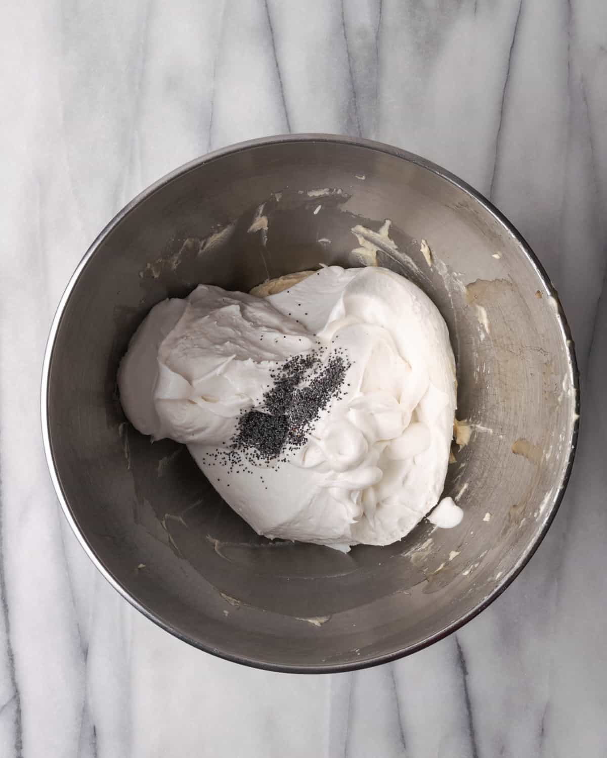 Whipped cream and poppy seeds added to a metal mixing bowl with cream cheese.