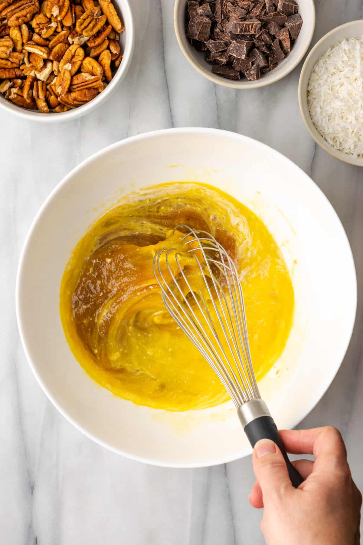 Eggs, sugar, corn syrup, vanilla, salt, and coconut milk being whisked together in a large white bowl.