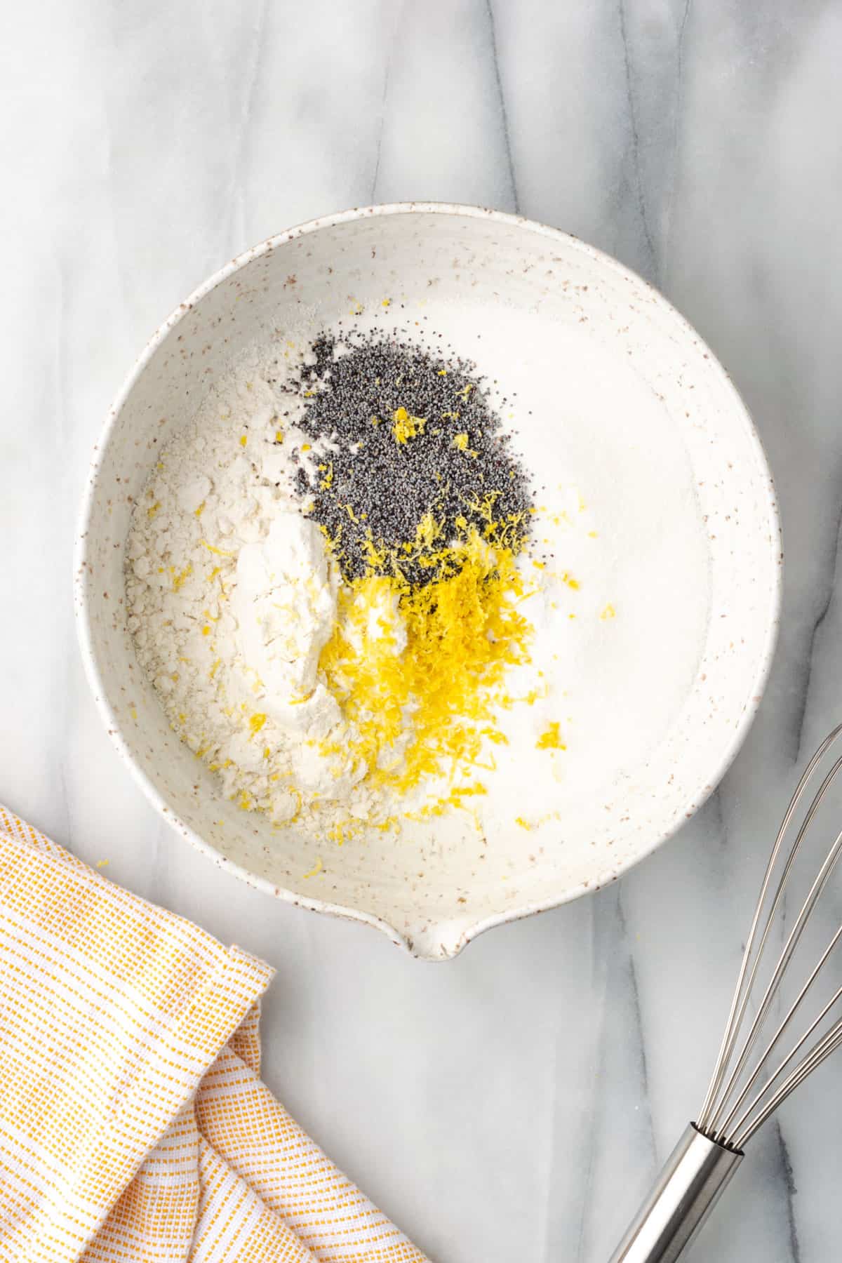 Flour, sugar, baking powder, salt, poppy seeds and lemon zest combined together in a large white bowl.