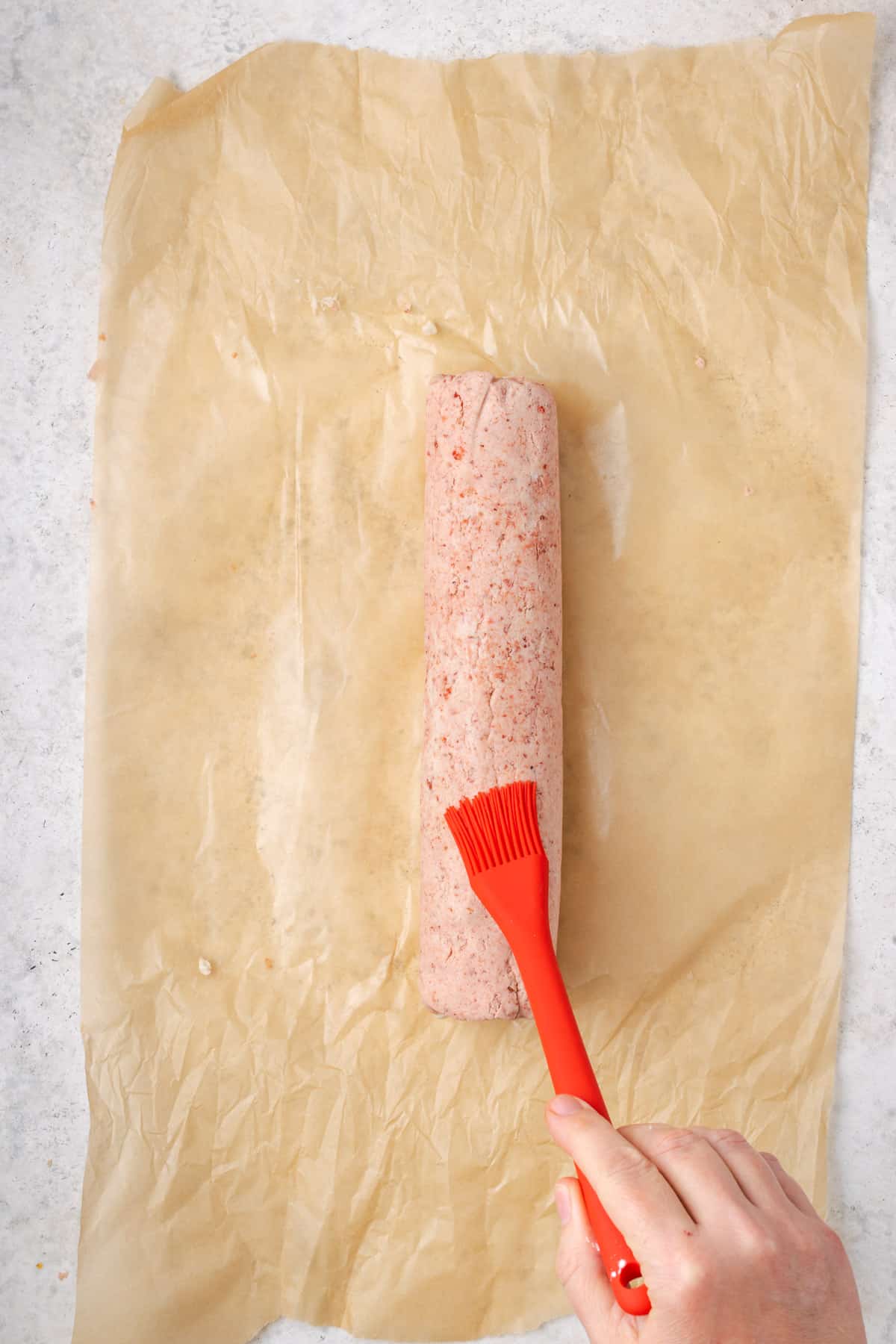 Log of strawberry cheesecake dough being brushed with water on a pastry brush.