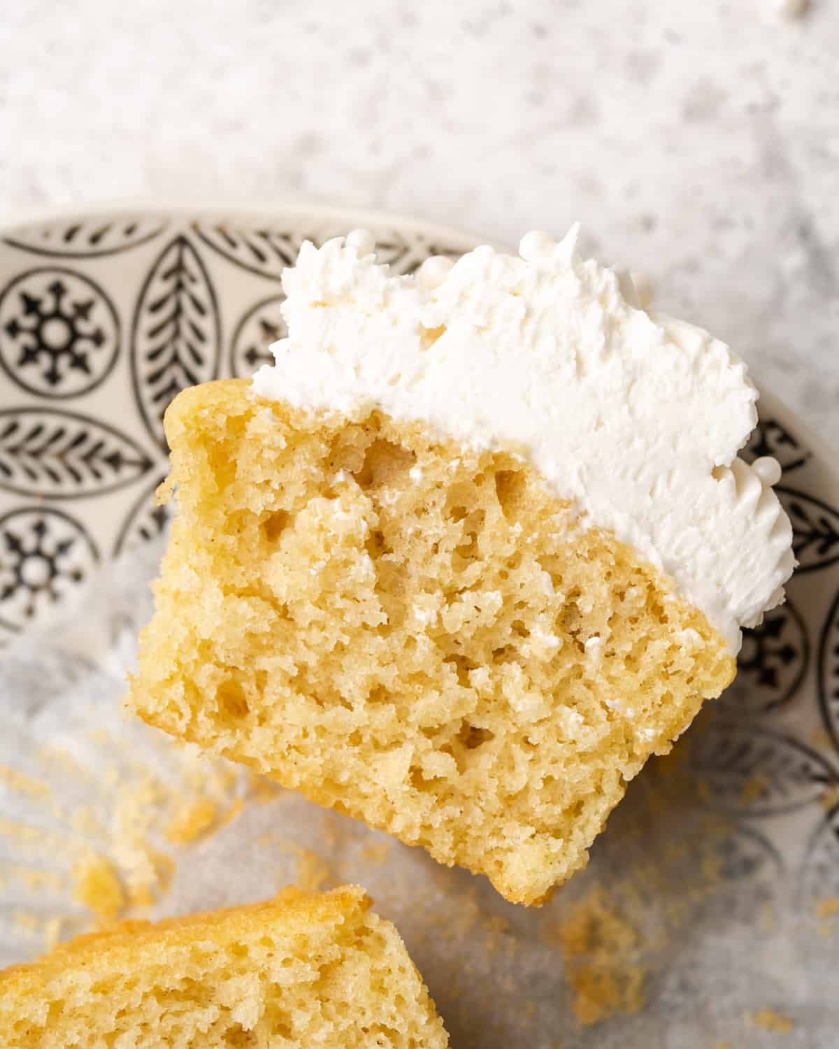 Sliced gluten free vanilla cupcake topped with vanilla frosting on a black and white print plate.