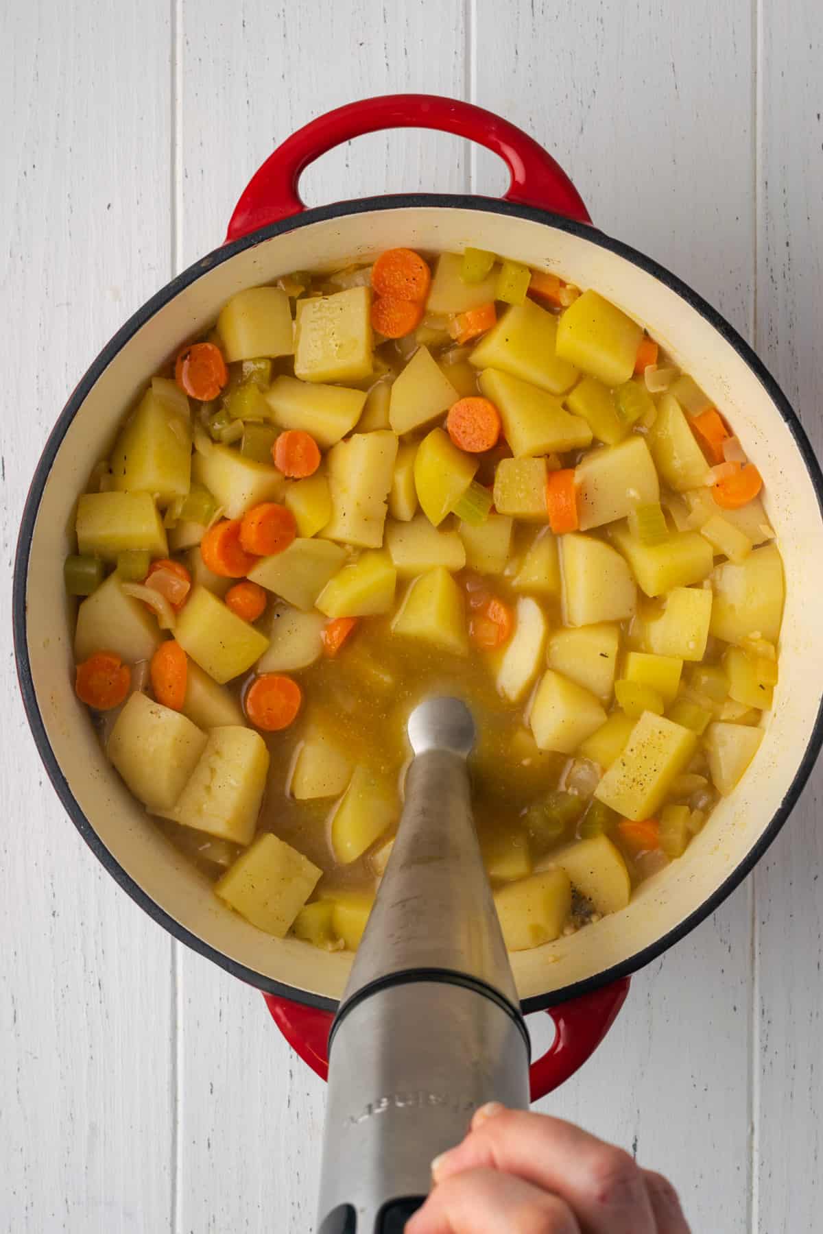 An immersion blender partially submerged in a pot of cooked potatoes and carrots.