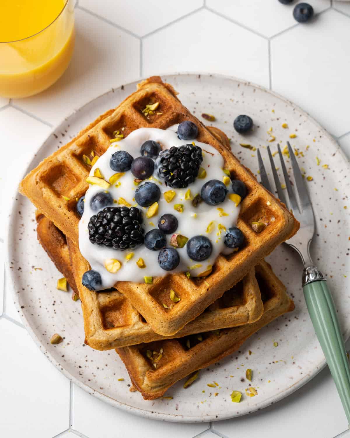 A stack of three golden oat flour waffles topped with dairy free yogurt, blueberries, blackberries and chopped pistachios.