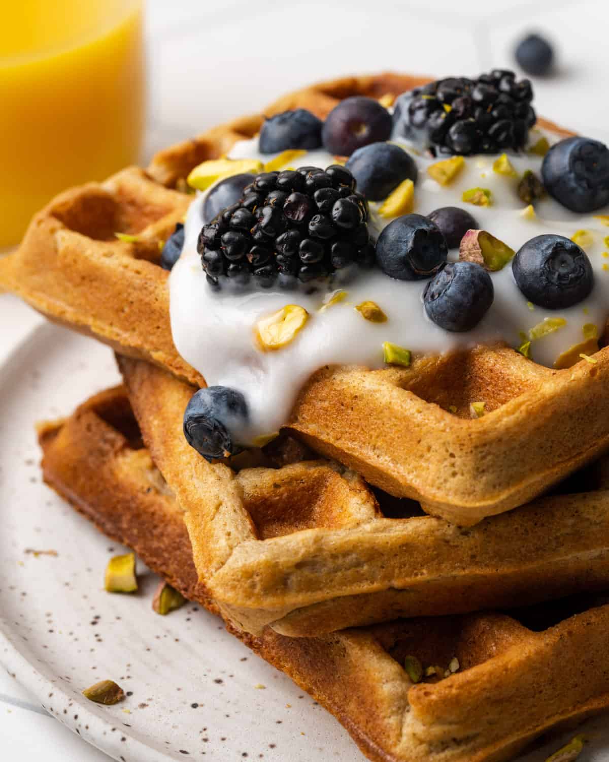 A closeup of a stack of three oat flour waffles topped with yogurt, blueberries and chopped pistachios.