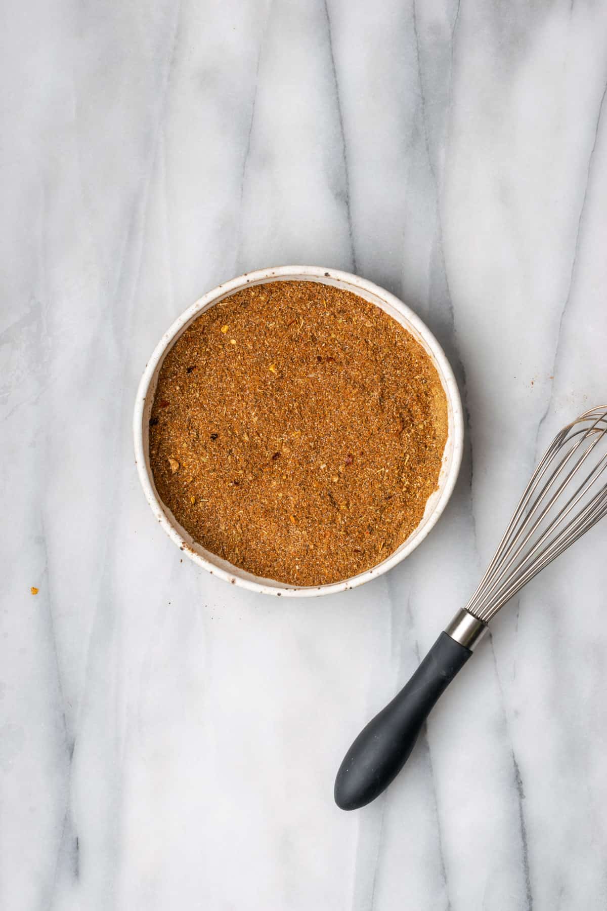 A bowl of mixed taco seasoning on a white table next to a whisk.