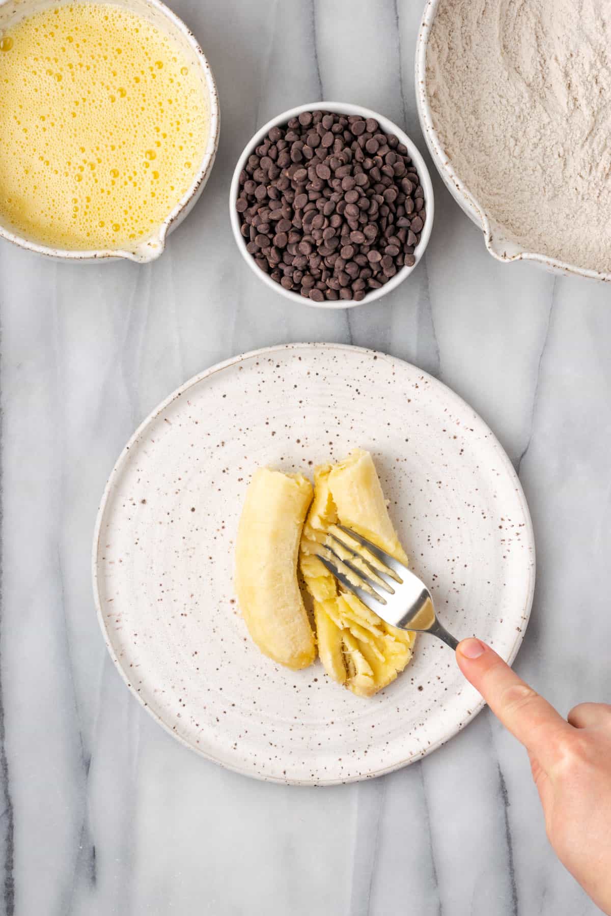 A banana being mashed on a plate with the tines of a fork.