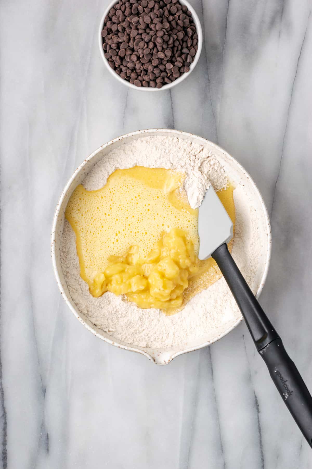 Egg mixture and mashed banana being folded into the dry ingredients in a large white bowl.