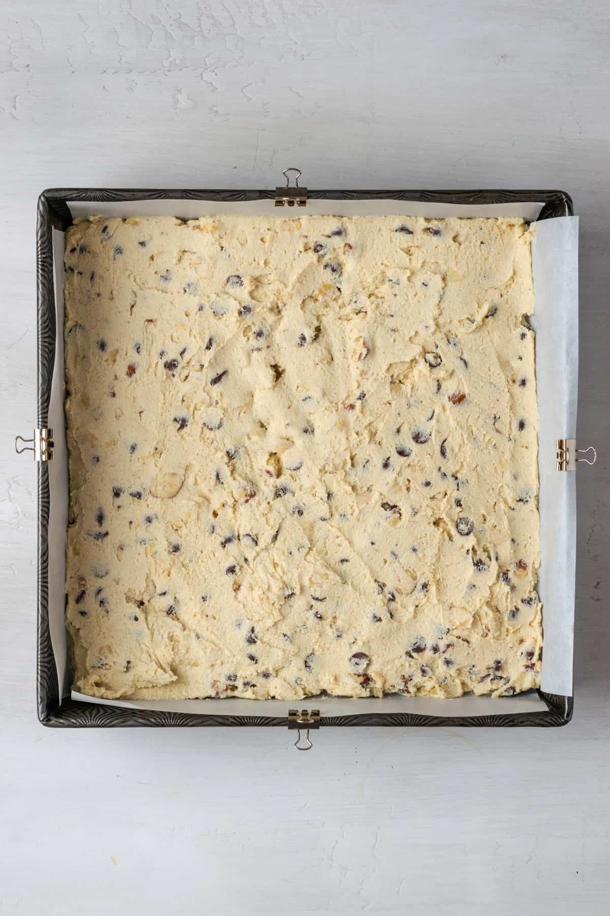 A square pan lined with parchment and filled with a layer of chocolate chip cookie dough.