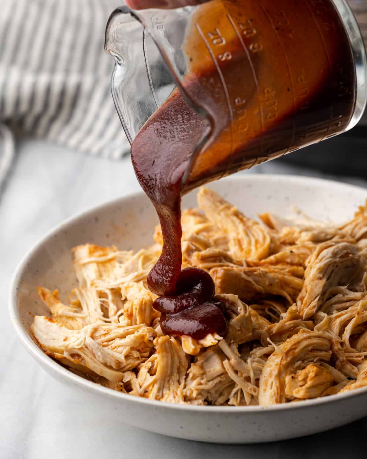 BBQ sauce being poured over pulled chicken in a serving dish.