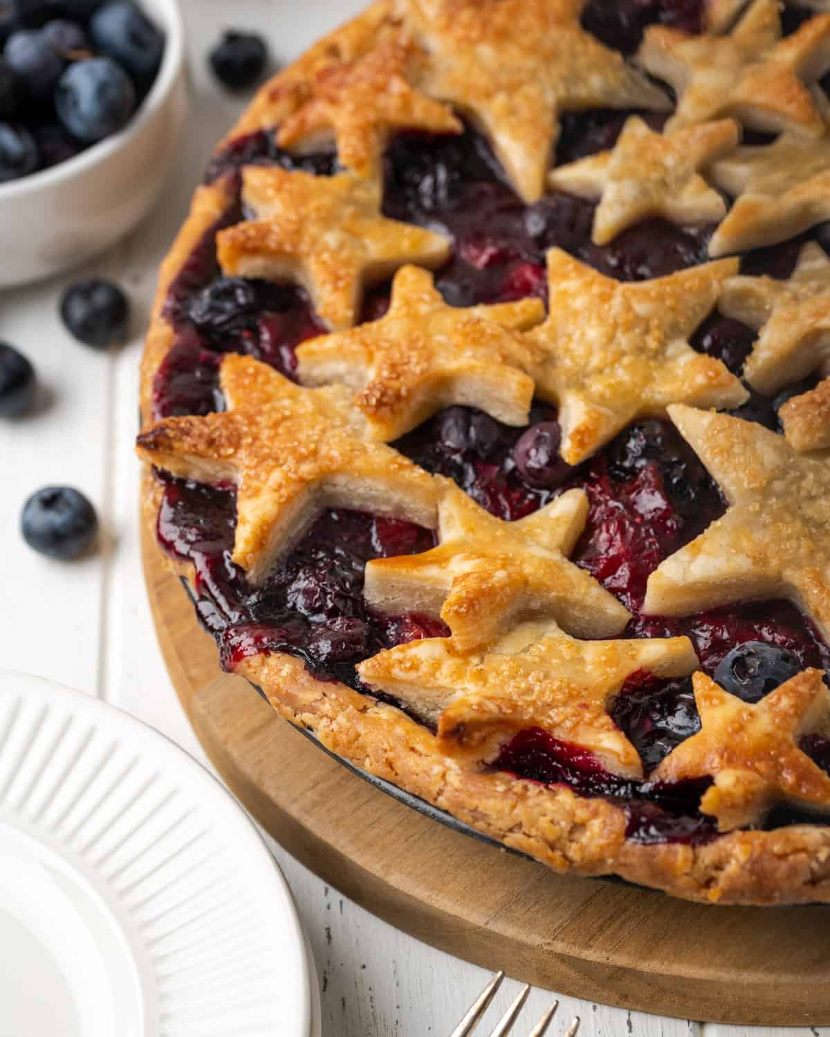 A freshly baked strawberry blueberry pie on a white table next to a bowl of fresh blueberries.