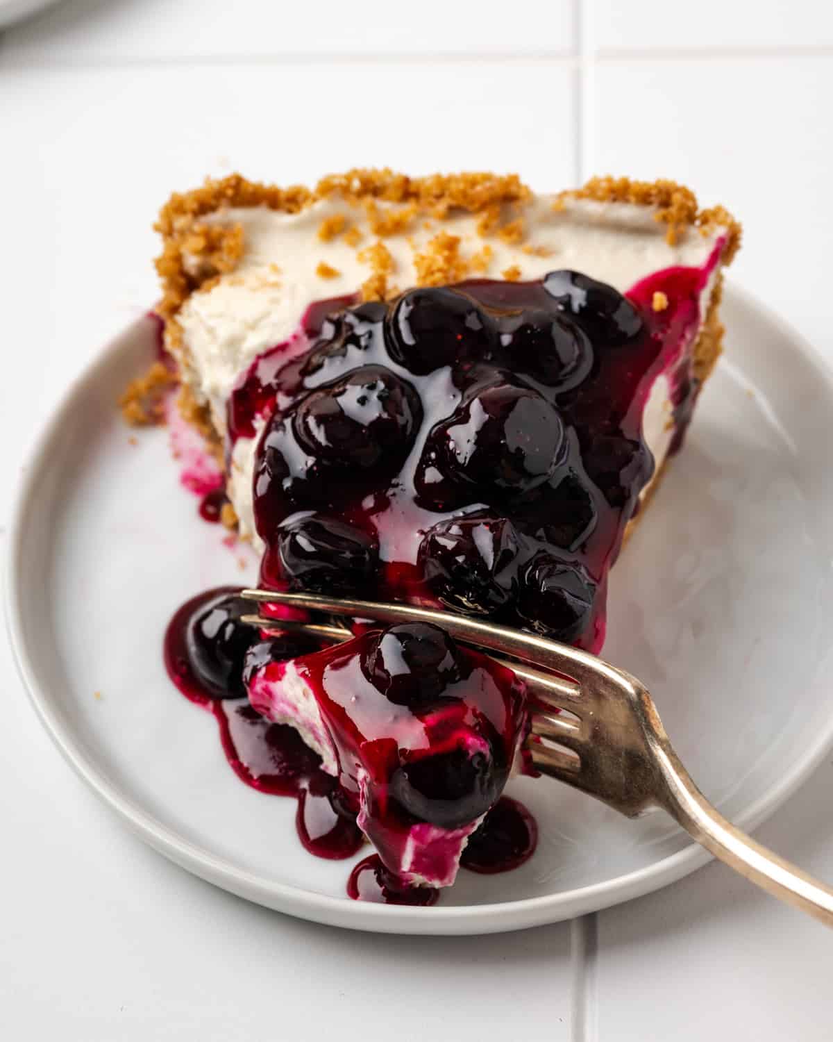 A bite being cut out of a slice of no bake blueberry cheesecake sitting on a white plate.