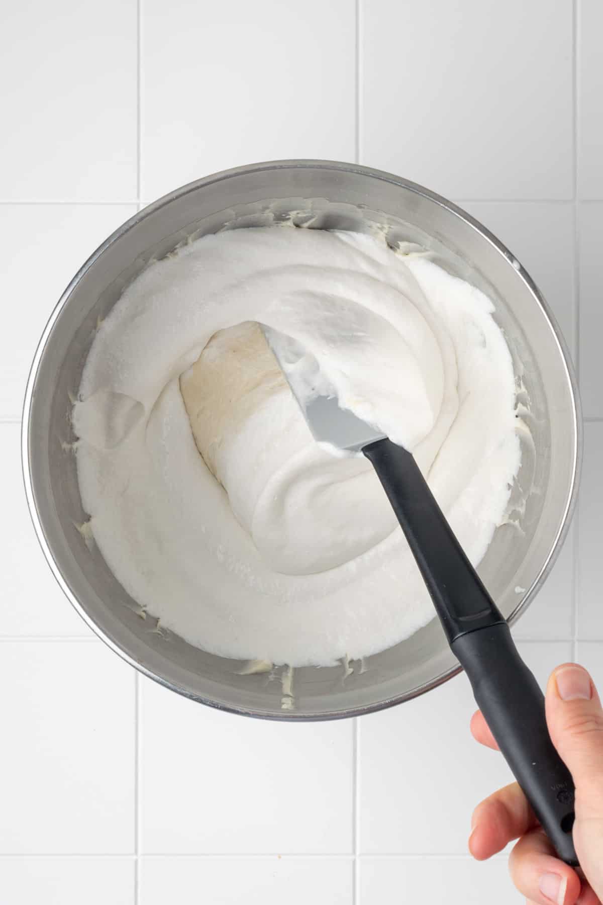 Whipped cream being folded into the cream cheese mixture in a metal mixing bowl.