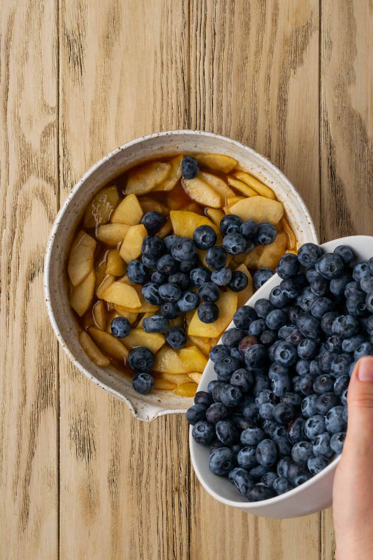 Fresh blueberries being poured into a bowl of cooked apples.