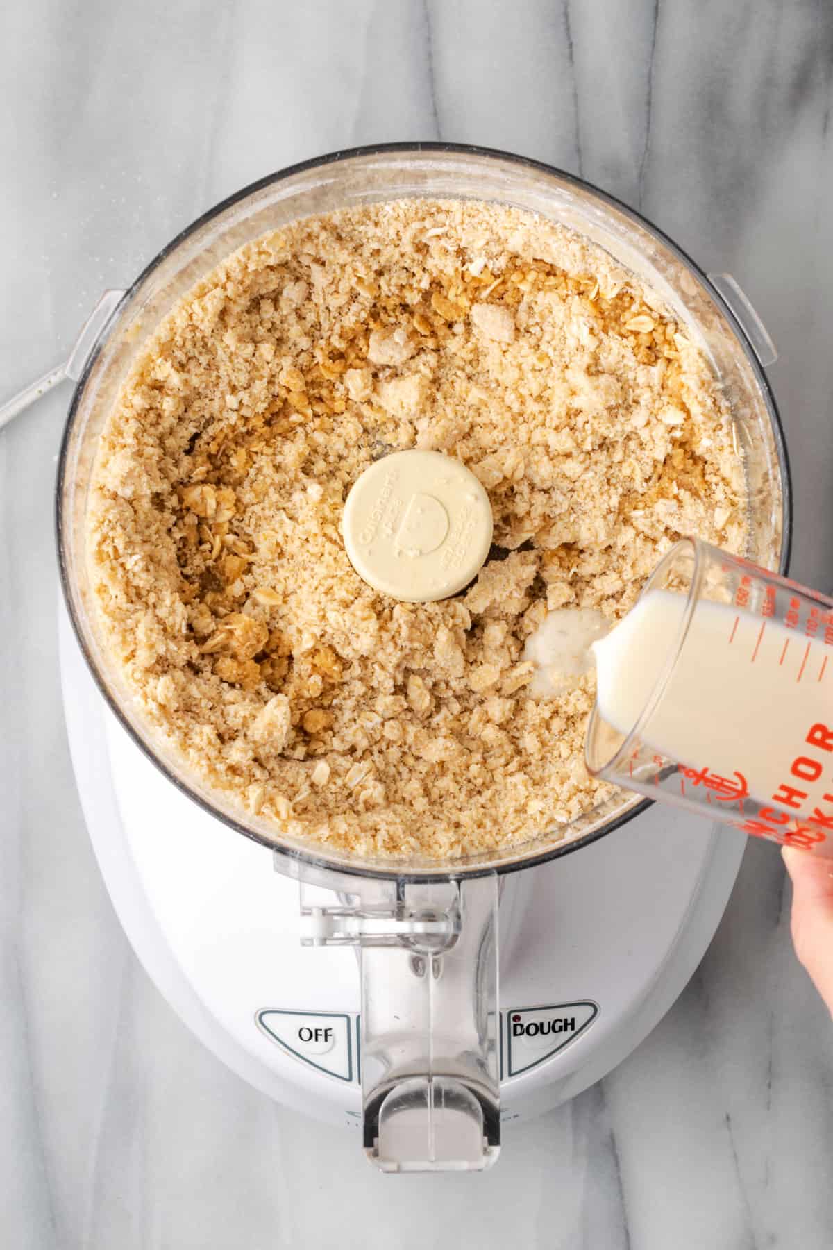 Milk being poured into the partially mixed crust dough in a food processor.