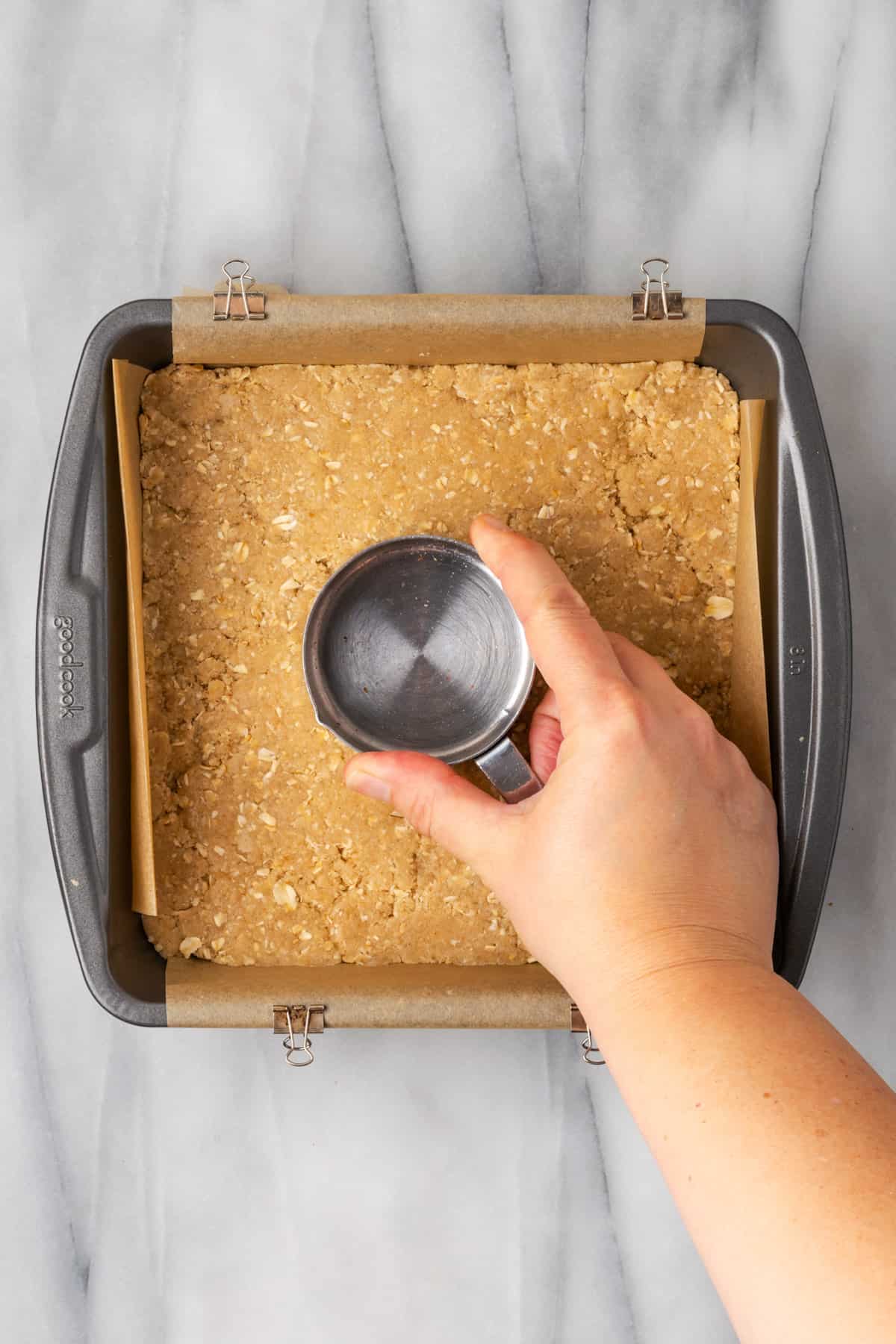 Dough being pressed into the bottom of a square pan with a metal measuring cup. 