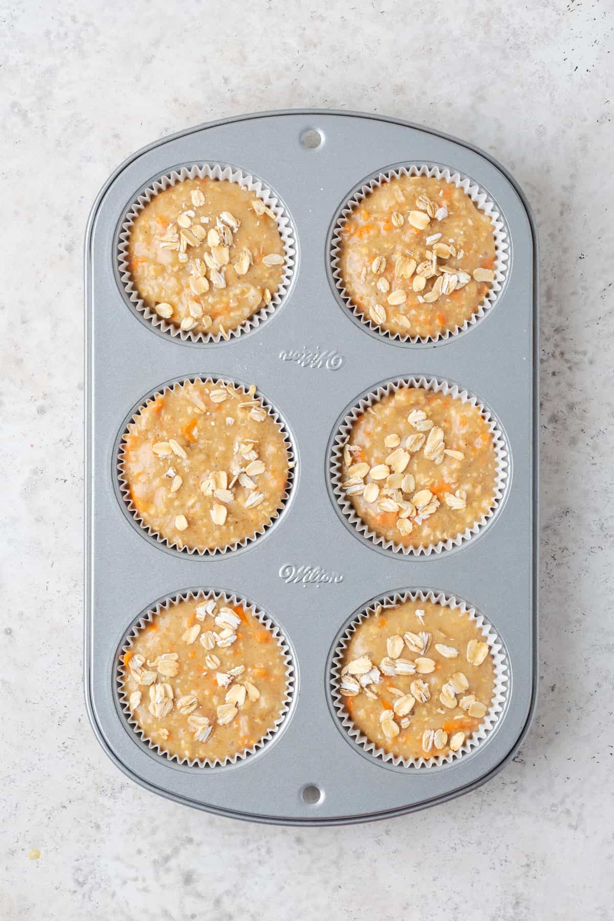 Unbaked applesauce carrot muffin batter topped with a sprinkle of oats in a 6 portion muffin tin.