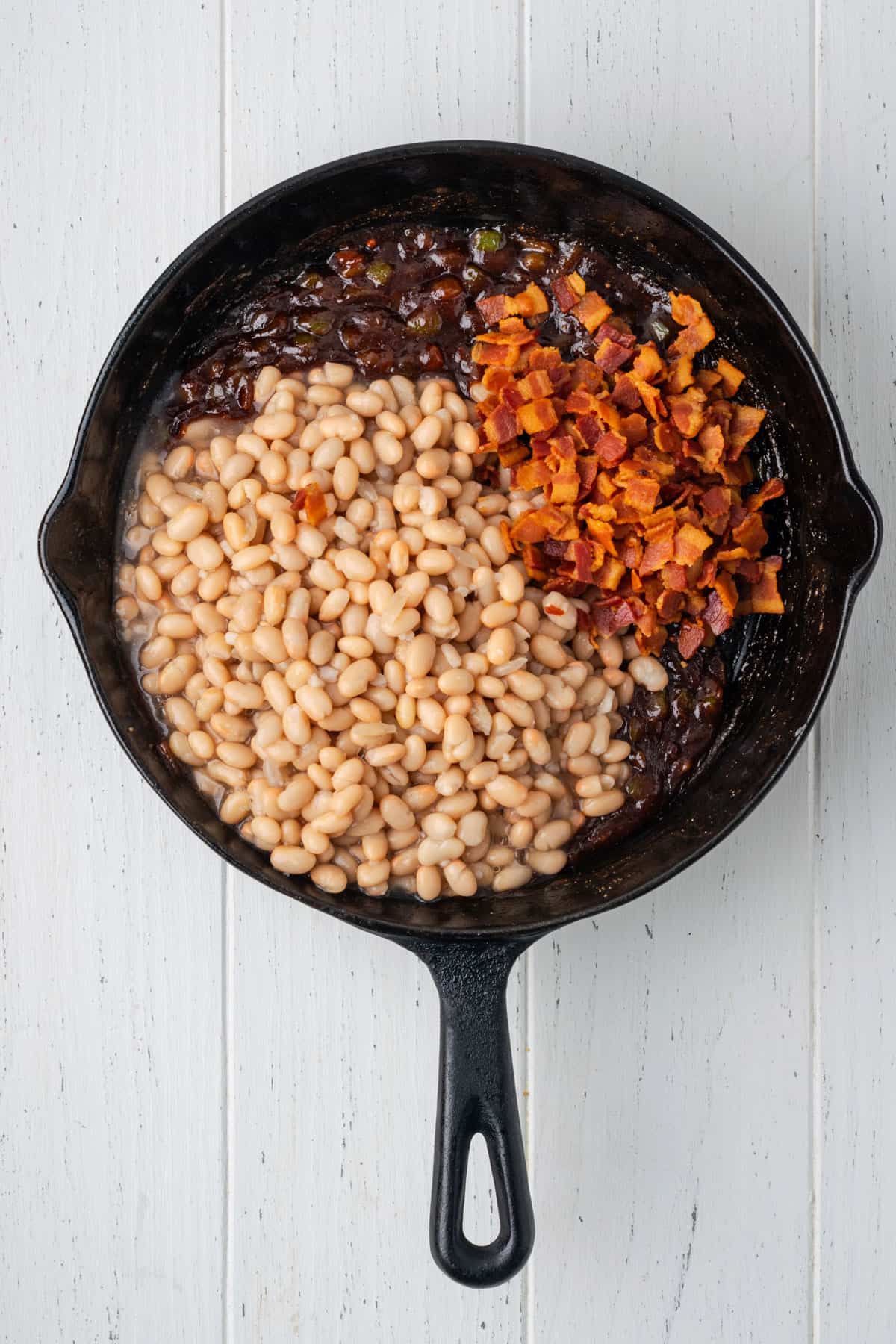 Navy beans and cooked bacon added to the bbq sauce in a cast iron skillet.