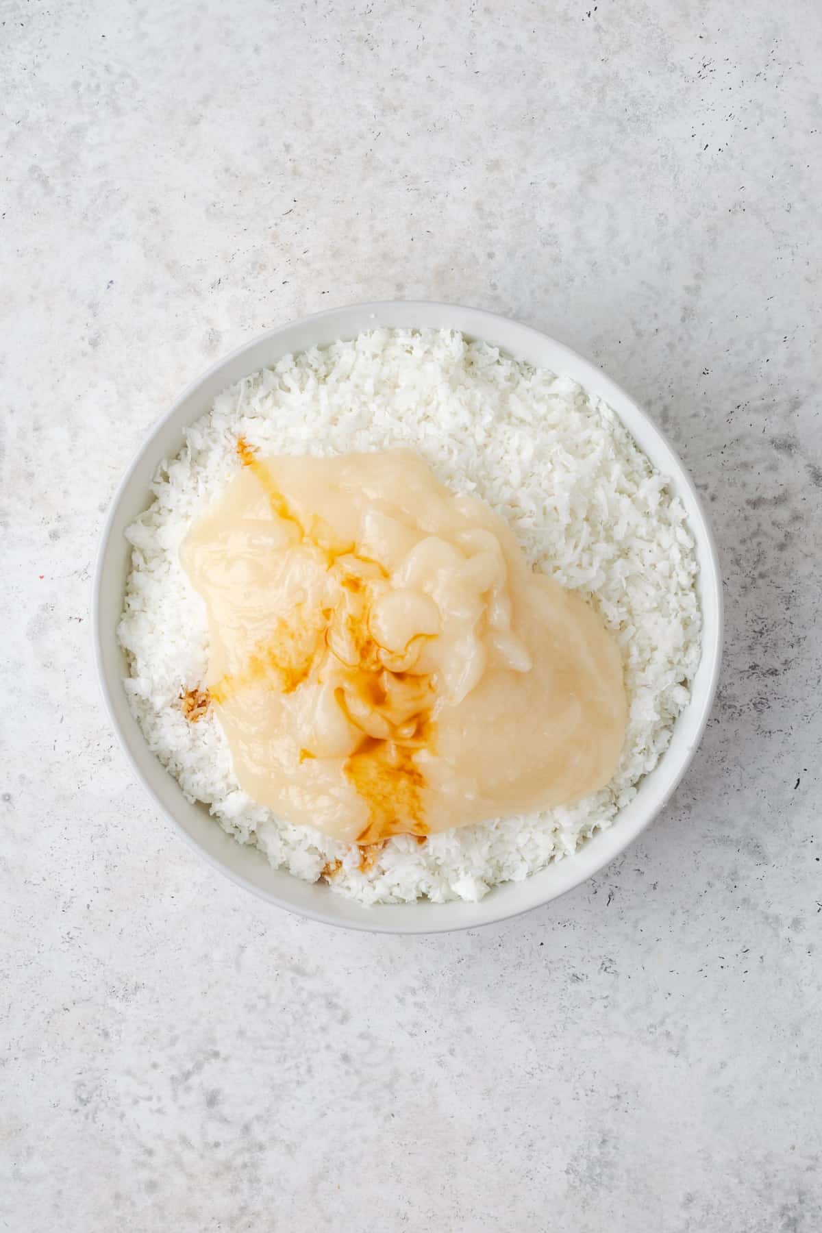 Shredded coconut, condensed milk and vanilla in a white mixing bowl. 