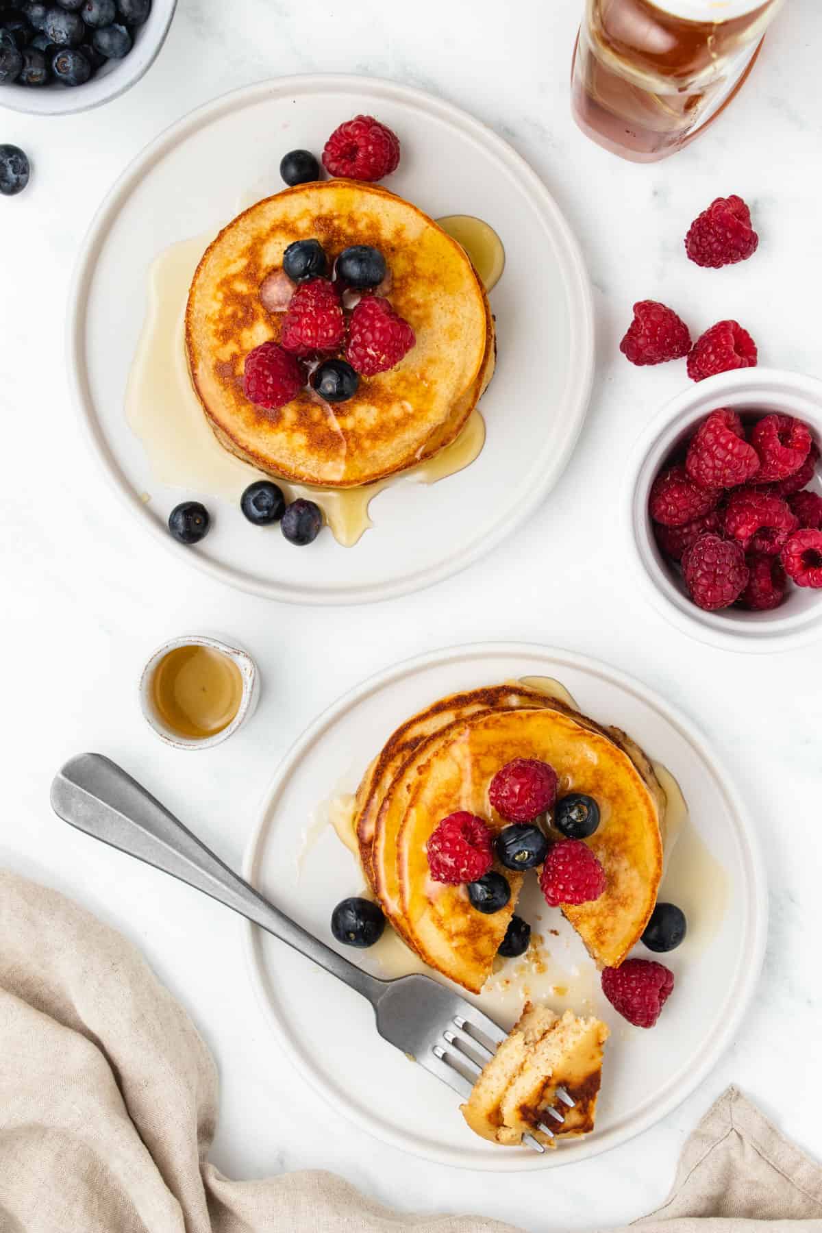 Two plates of grain free pancakes topped with maple syrup and fresh berries on a white table.