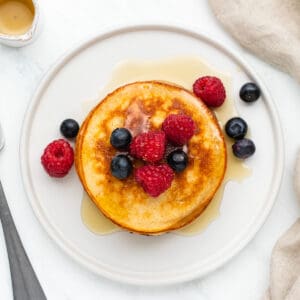 A stack of grain free pancakes on a white plate, topped with syrup and fresh berries.