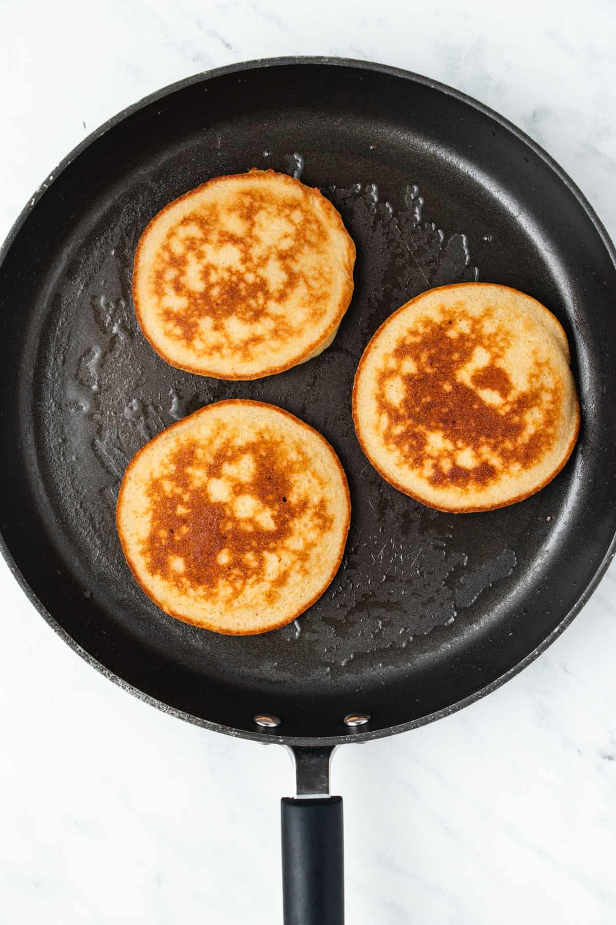 Three pancakes cooking in a black frying pan. 