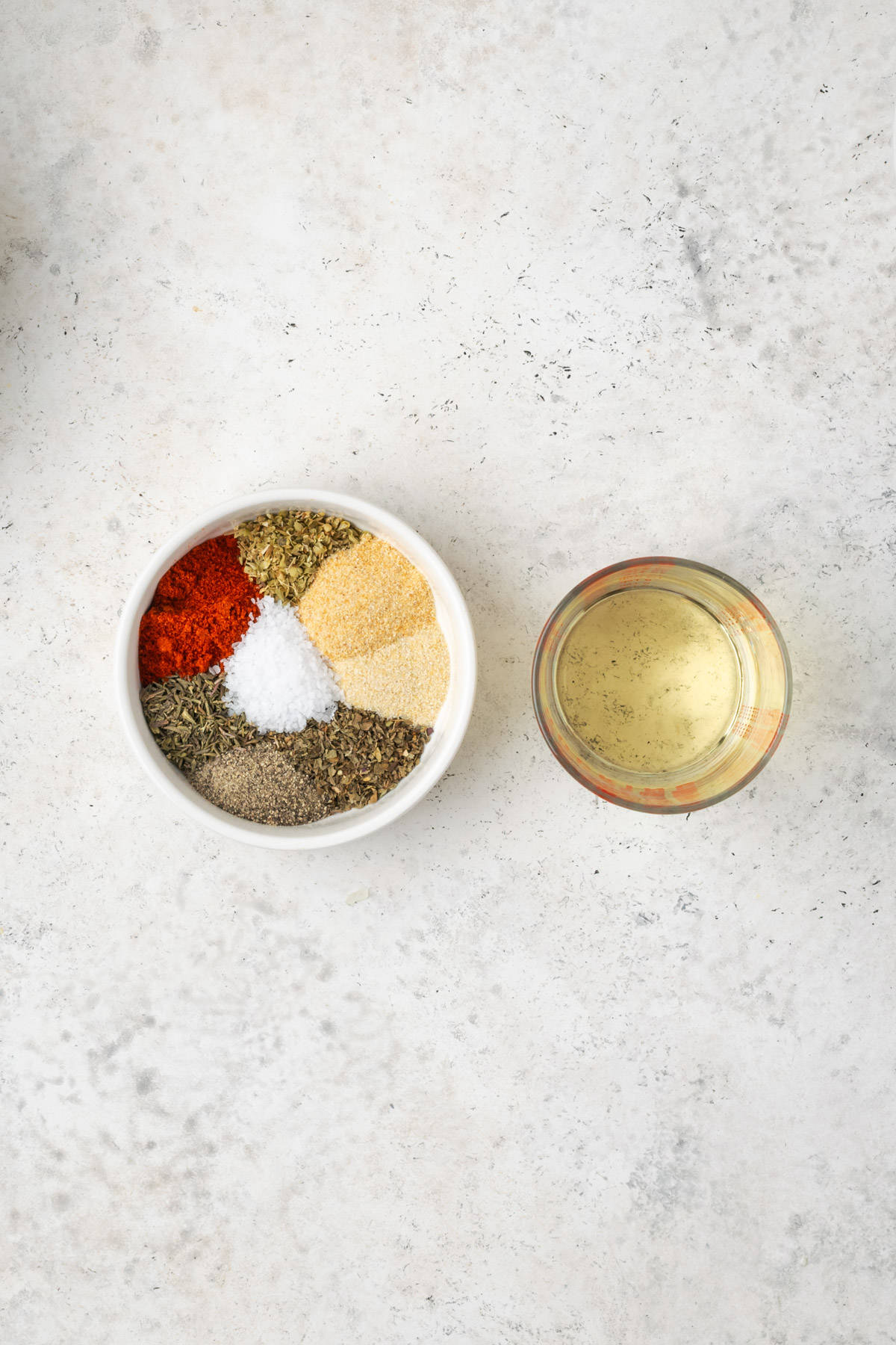 A small bowl of spices and a measuring cup with olive oil sitting on a white table.