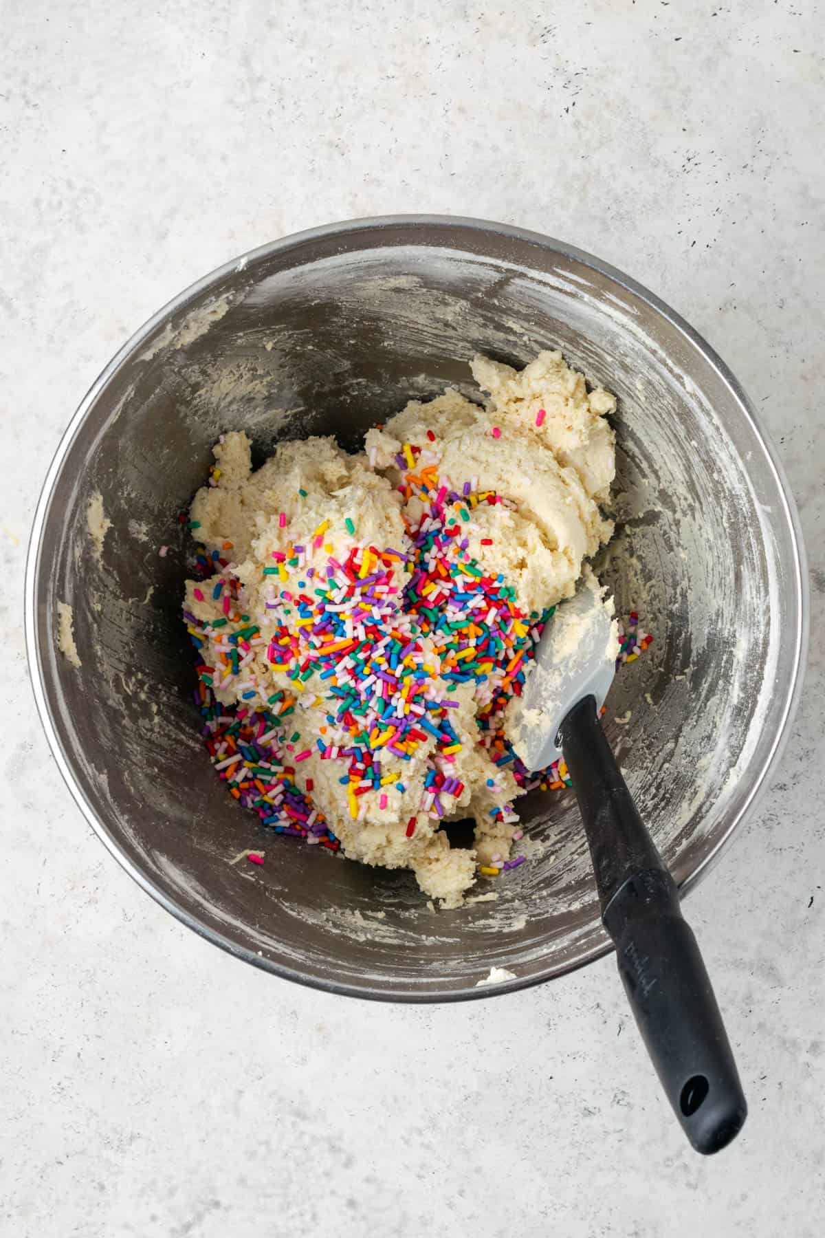 Rainbow sprinkles being folded into the sugar cookie dough with a spatula.