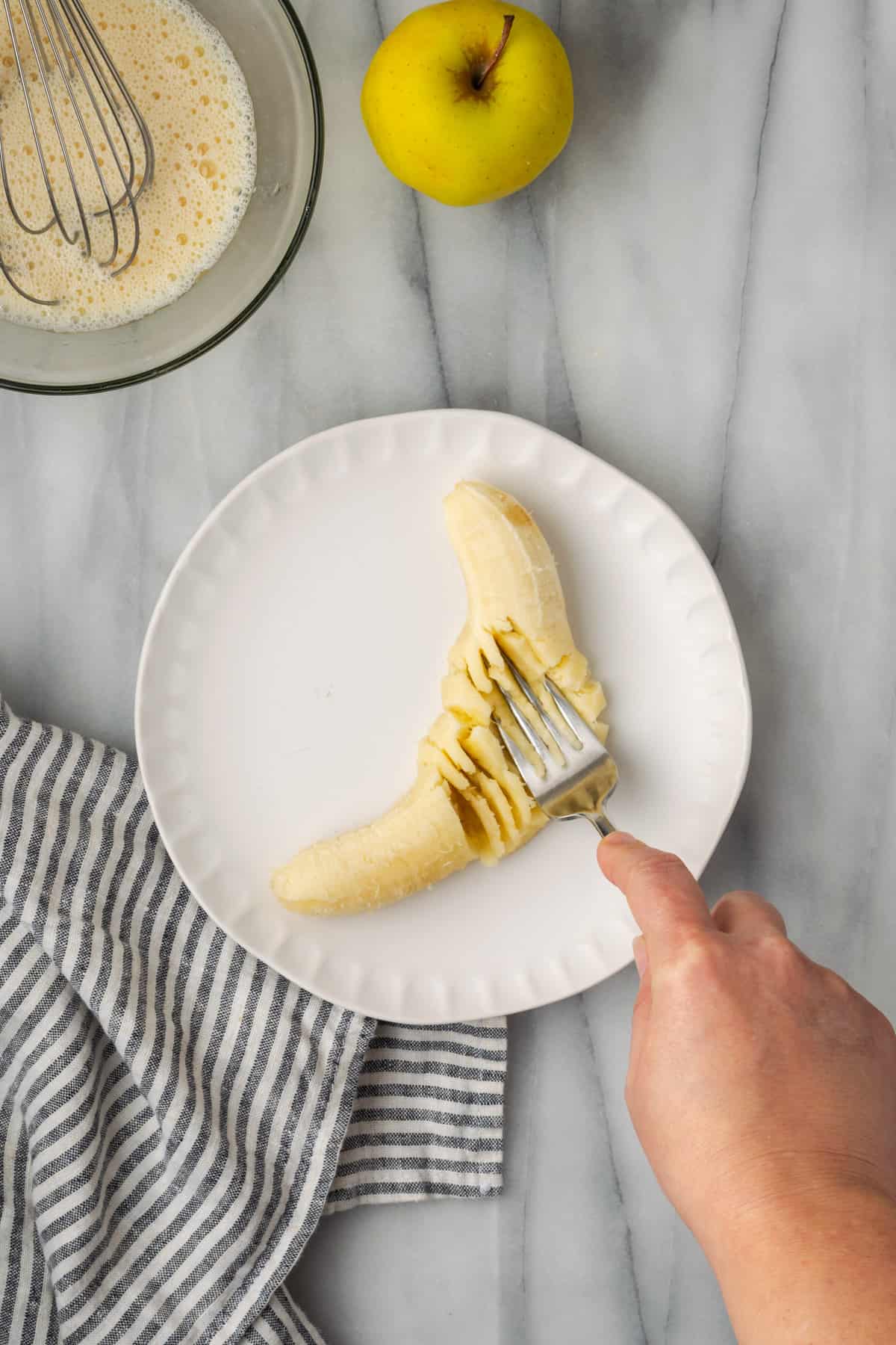 A single banana being mashed with a fork on a white plate.