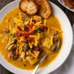A bowl of hamburger potato soup topped with shredded cheese and bacon and served along side toasted bread.