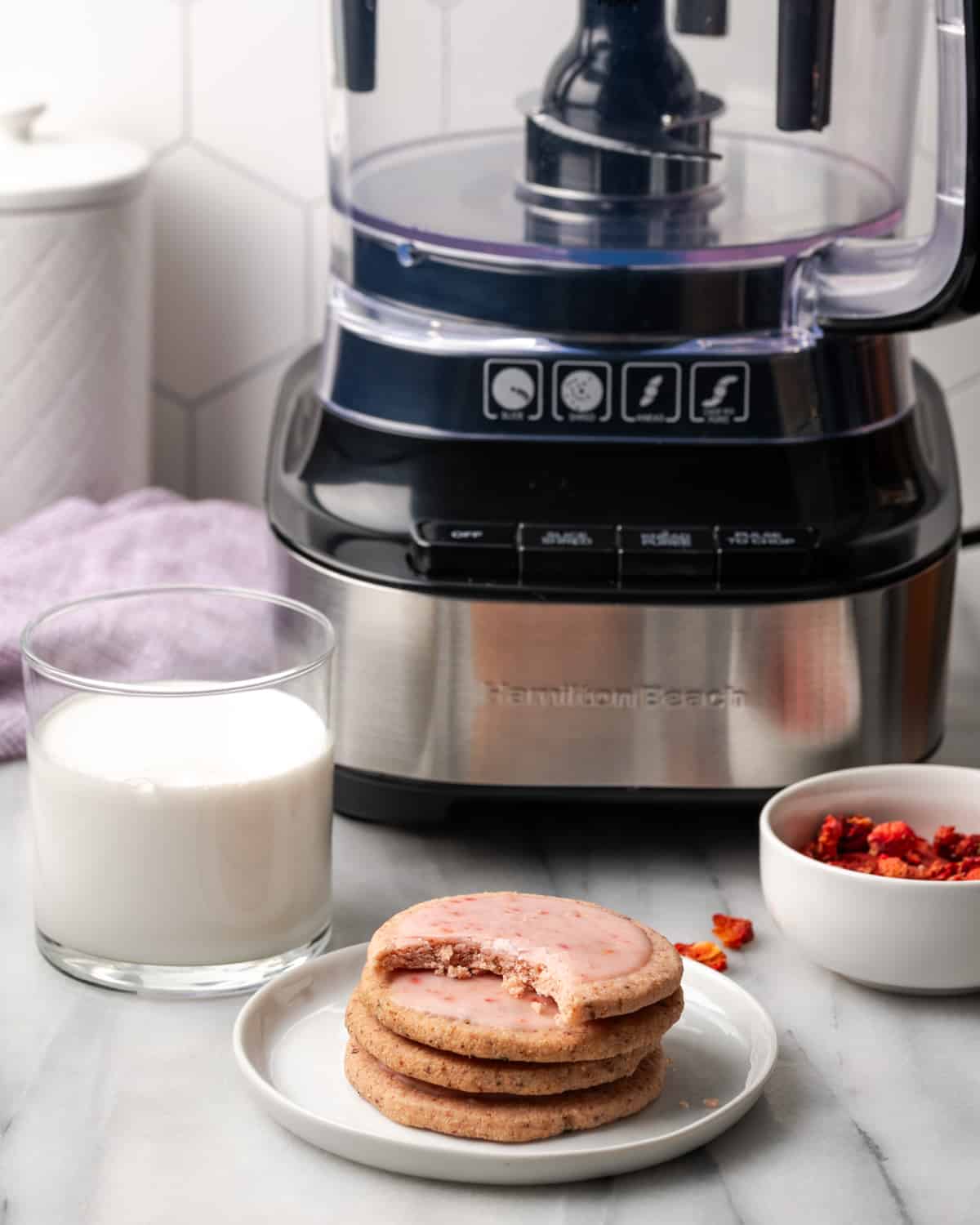 A stack of strawberry shortbread cookies sitting next to a food processor and glass of milk.