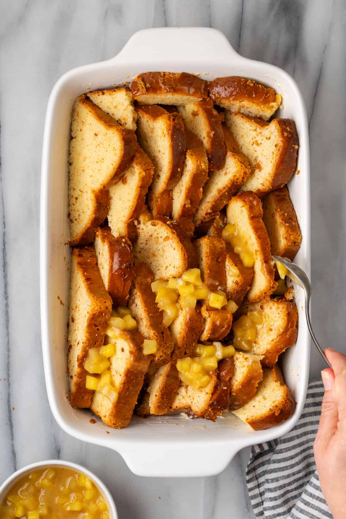 A baking dish full of soaked slices of brioche bread being topped with spoonfuls of apple pie filling. 