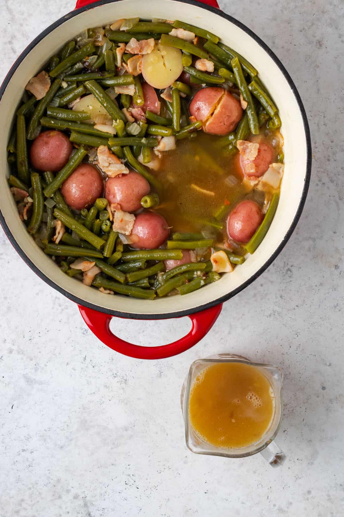 A pot of cooked green beans and potatoes next to a measuring cup filled with pot likker.